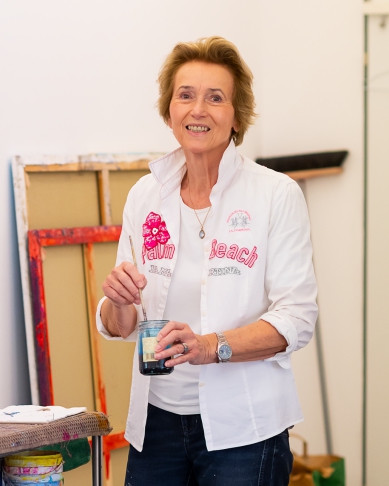 A smiling older woman stands in an art studio, holding a paintbrush and a small container of paint. She wears a white shirt with "Palm Beach" written on it, and several colorful canvases are visible in the background.