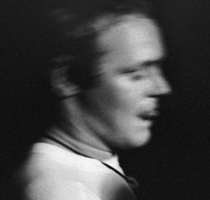 A black-and-white photo of a person in motion, with their face and features blurred, wearing a light-colored shirt and looking to the side against a dark background.