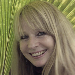A woman with long blonde hair smiles in front of a large, green palm leaf that forms the background.