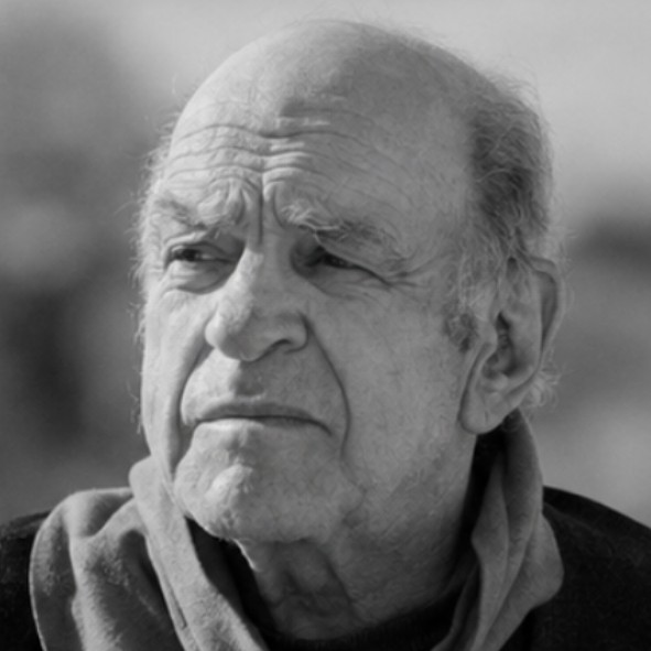 An elderly man with a bald head and gray hair on the sides wears a scarf and looks thoughtfully into the distance. The photo is in black and white.