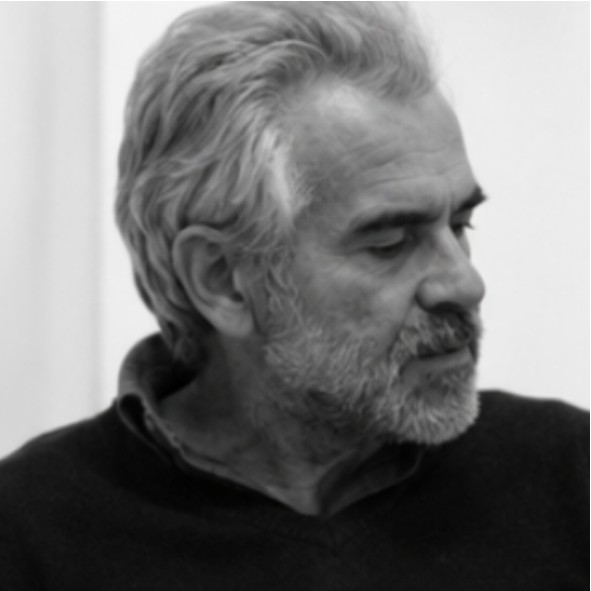 Black and white photo of an older man with gray hair and a beard, looking to his left. He is wearing a dark sweater over a collared shirt and appears thoughtful. The background is plain and light-colored.
