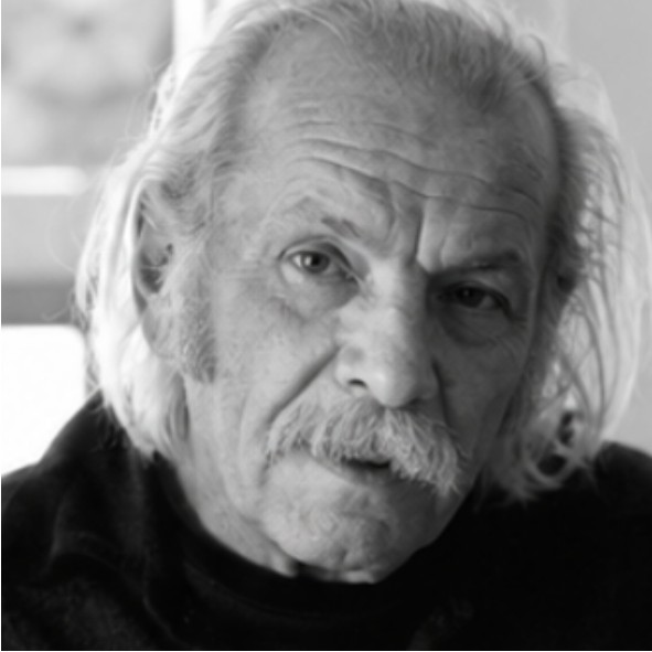An elderly man with long white hair and a mustache looks into the camera with a serious expression. The image is in black and white.