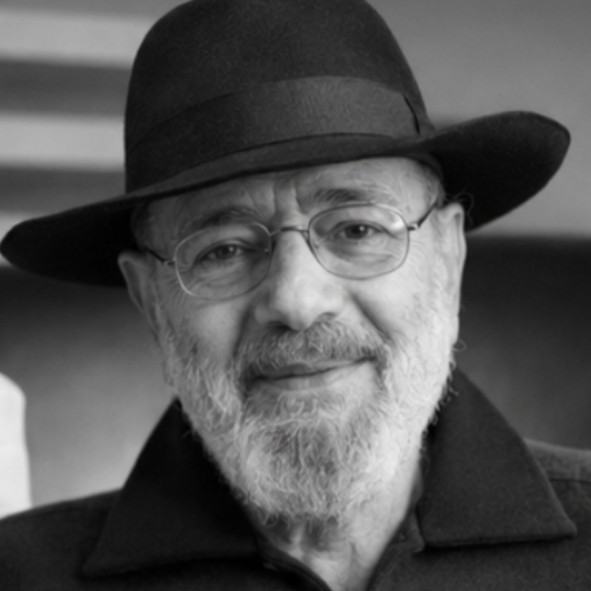 An elderly man with a beard and glasses smiles gently at the camera. He is wearing a dark hat and a dark collared jacket. The image is in black and white.