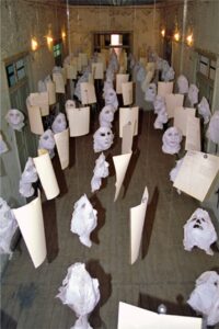 An art installation in a long room features ghostly white masks and sheets of paper suspended from the ceiling with string, creating an eerie and surreal atmosphere. The walls and floor appear old and worn.