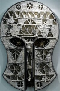 A textured, abstract sculpture shaped like a stylized human face or mask, featuring geometric patterns, triangles, and metallic accents in gray, white, and black tones. The eyes are round and slightly recessed.