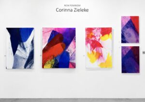 Five colorful abstract paintings by Corinna Zieleke are displayed on a white gallery wall under the title "New Feminism." The artworks feature vibrant reds, blues, purples, and yellows with dynamic shapes.