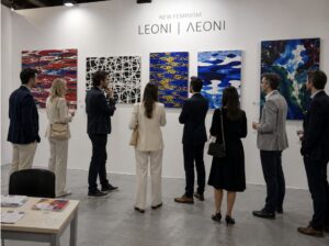 A group of people stand in front of a gallery wall displaying colorful abstract paintings, engaging in conversation. The wall features the text "NEW FEMINISM LEONI | ΛΕΟΝΙ" above the artworks.