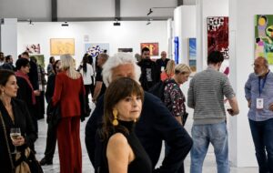 People walk around and observe colorful artwork displayed on white walls at a busy art gallery or exhibition. Some individuals are engaged in conversation, while others examine the art. The atmosphere appears lively and social.