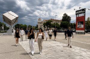 A group of people walk through a city square near the MOMus Contemporary Art Museum under a cloudy sky, with a large abstract sculpture and historic building in the background.