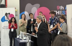 A man speaks at a podium during the 9th Art Thessaloniki art fair, while a group of people stand behind him clapping. Colorful event branding is visible in the background.