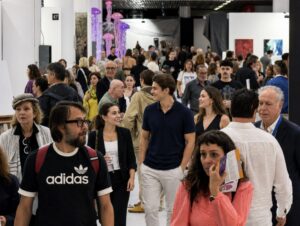 A large crowd of people walks through a busy indoor art exhibition, with various artworks displayed and bright purple sculptures in the background. Most people appear engaged, talking or observing their surroundings.