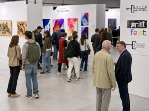 People walk and converse in a contemporary art exhibition with colorful abstract paintings and graphic prints displayed on white walls; signage reads “print fest” and “Corinna Zeleke.”.