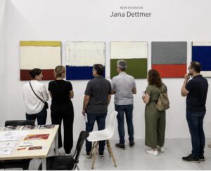 Six people stand in front of a white wall displaying abstract paintings by Jana Dettmer. The group observes the colorful, geometric artworks arranged in a row, under the sign "NEW FEMINISM Jana Dettmer.