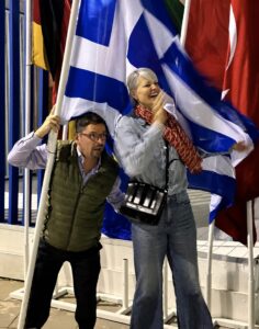 Two people enthusiastically wave a large Greek flag among other national flags. The woman is smiling and cheering, while the man poses beside her, both dressed in casual clothing.