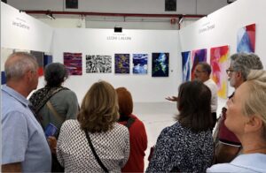 A group of people stands in a white art gallery, attentively listening to a man speak about the colorful abstract paintings displayed on the walls. The atmosphere is engaged and focused.