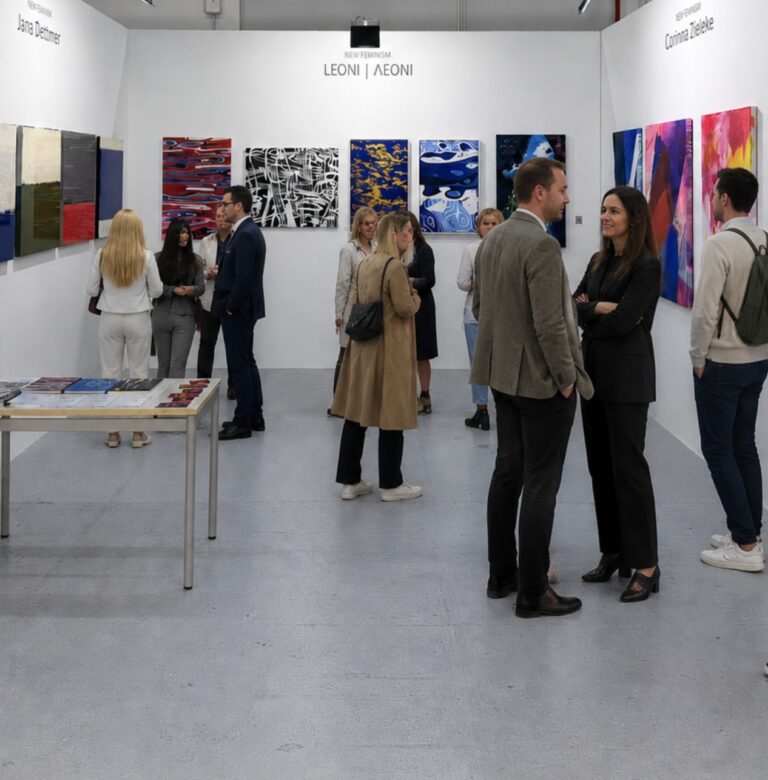 People interact and view abstract paintings at a modern art gallery exhibition. Groups are engaged in conversation, and colorful artworks are displayed on white walls in a bright, open space.