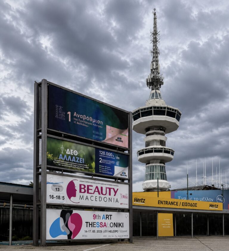 A cloudy sky above a tall, modern telecommunications tower. In the foreground, colorful event posters and signs in Greek and English are displayed on a board near an entrance with yellow signage.